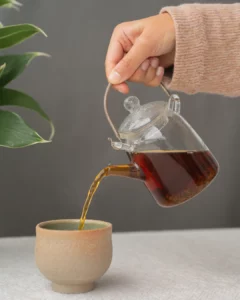 Hand schenkt zwarte Ceylon “UVA Highland” thee met cafeïne uit een glazen theepot in een kopje.